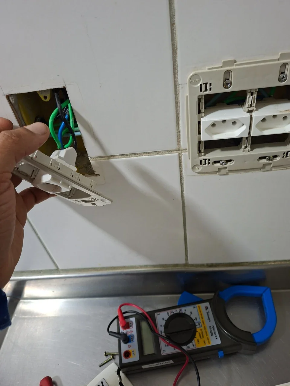 Tomada residencial aberta durante avaliação elétrica, com fiação visível e multímetro de alicate.